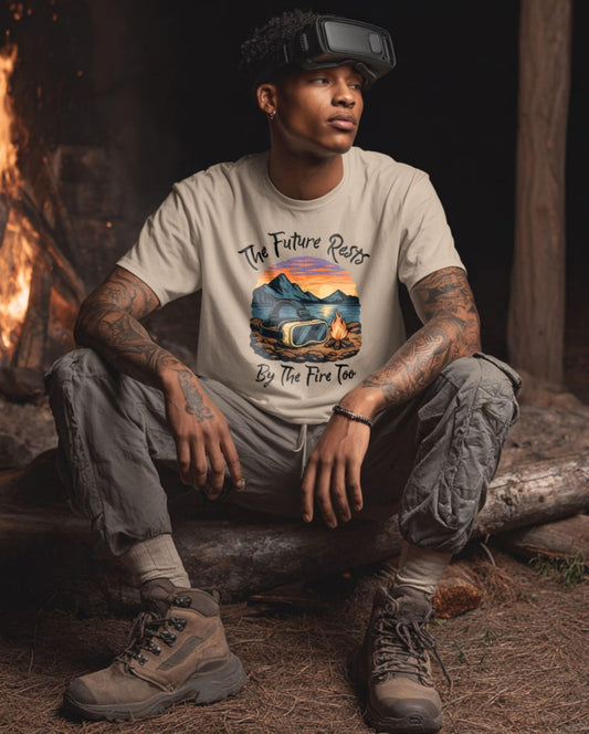 male model wearing a sand-colored t-shirt with a graphic of a VR headset resting by a campfire at sunset, surrounded by mountains, with the text "The Future Rests By The Fire Too"